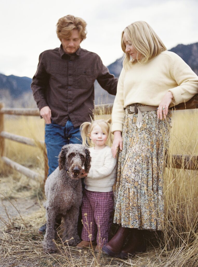 Family standing together with their dog in tall grass near a wooden fence with mountain scenery at South Boulder Peak Trailhead.
