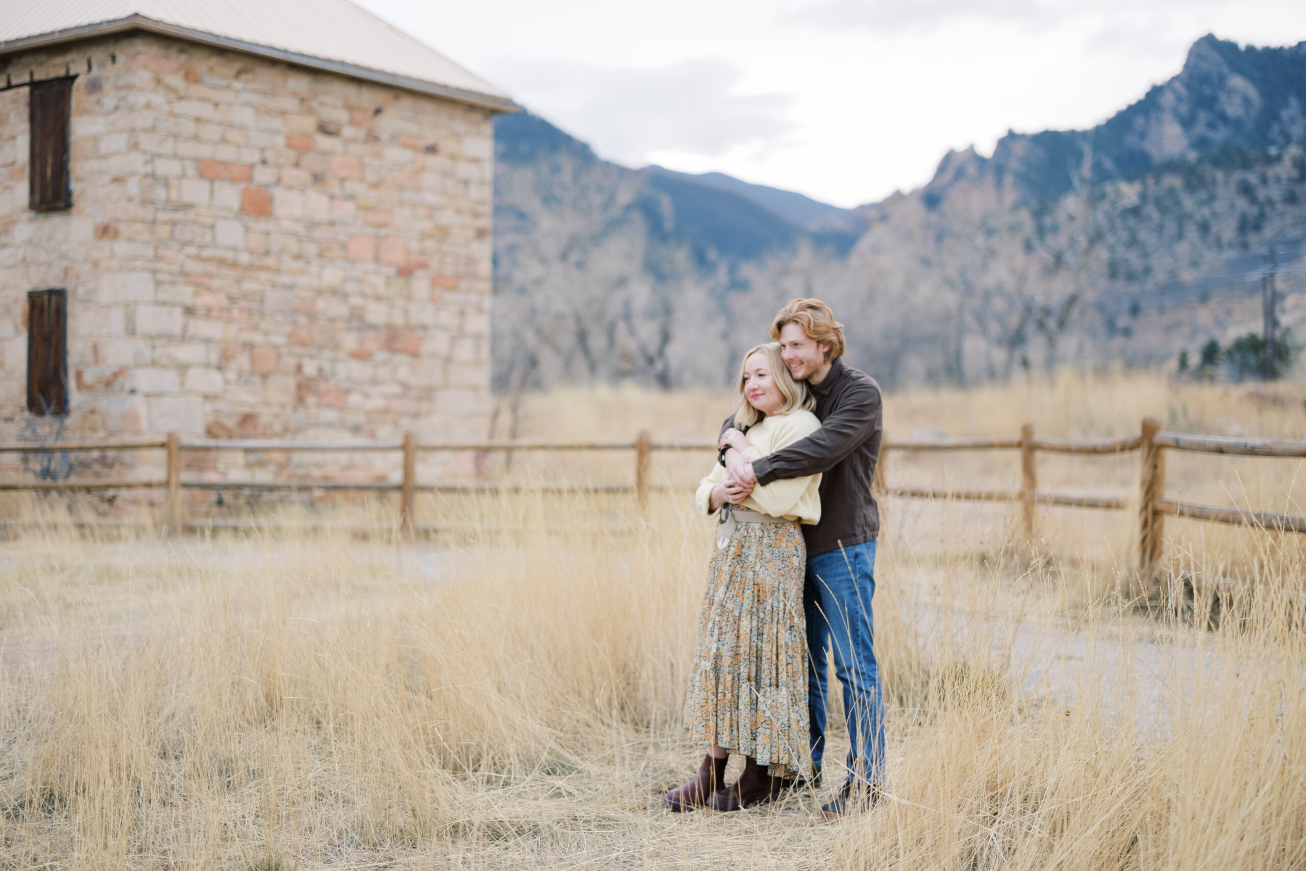 A couple embracing in tall golden grass near a rustic stone building with the Flatirons in the background at South Boulder Peak Trailhead.